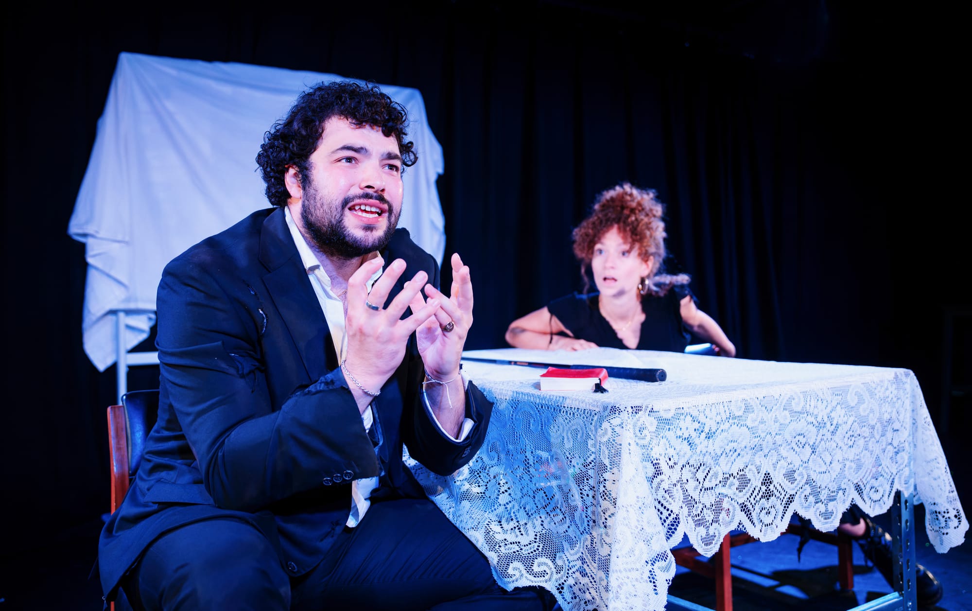 Dylan Corbett-Bader sits opposite Gemma Barnett at a table covered in white lace. Corbett-Bader makes an anguished expression and Barnett is leaning across the table to speak to him.