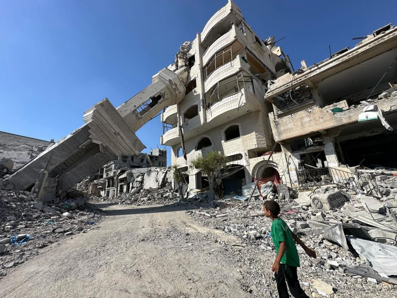 A small boy in a green shirt looks out at a destroyed neighbourhood. A tower has collapsed into the side of a building. There is rubble along the street where he stands. 