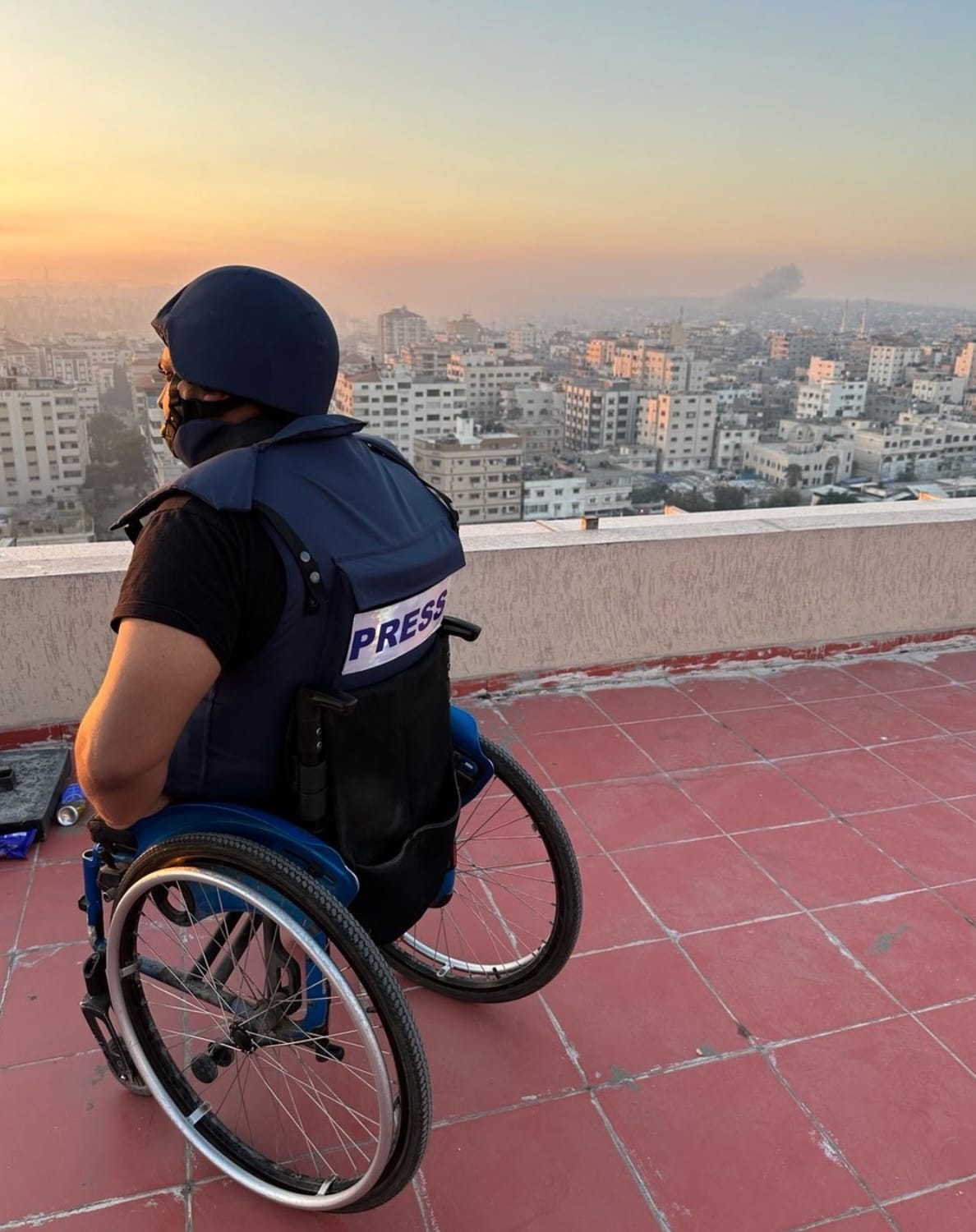 Momin Faiz is pictured in a press vest in his wheelchair on the rooftop of a building. He is looking out at the skyline of building. The sun is setting and smoke from an airstrike is rising in the distance. 