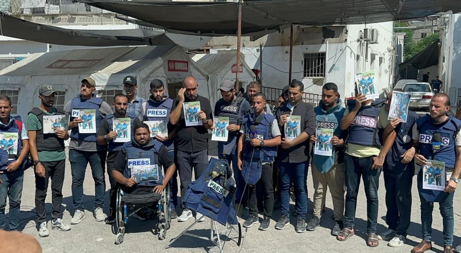 Seventeen men stand in a line, facing the camera and holding a piece of paper with the photos of their colleagues, Ismail and Rami, who were killed by an Israeli airstrike.