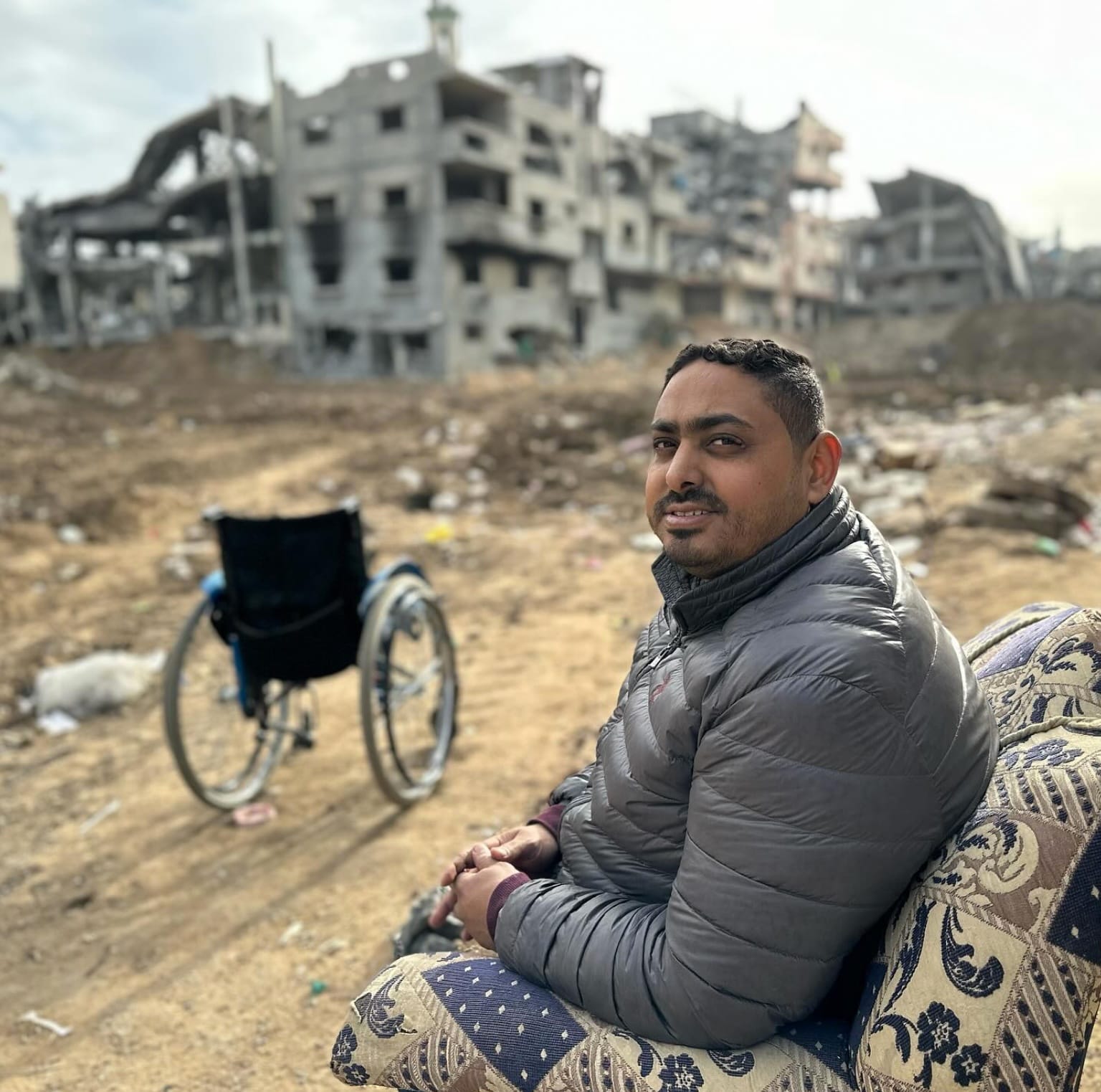 Momin sits in a patterned arm chair and looks at the camera. Behind him, his wheelchair is empty in front of a bombed building. 