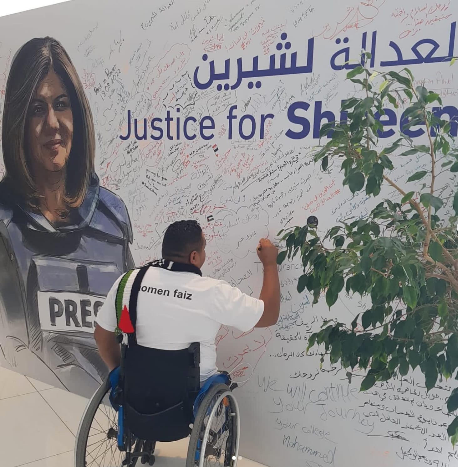 Momin is pictured signing a large white wall covered in other signatures and writing. The wall has a mural of Shireen Abu Akleh and says "Justice for Shireen". Momin is wearing a scarf with the Palestinian flag on it and he is in a wheelchair. 