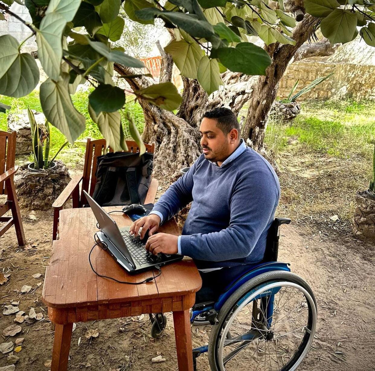 Momin Faiz sits in his wheelchair at a small, wooden table. He is typing on his laptop. The table is under a tree in a garden.