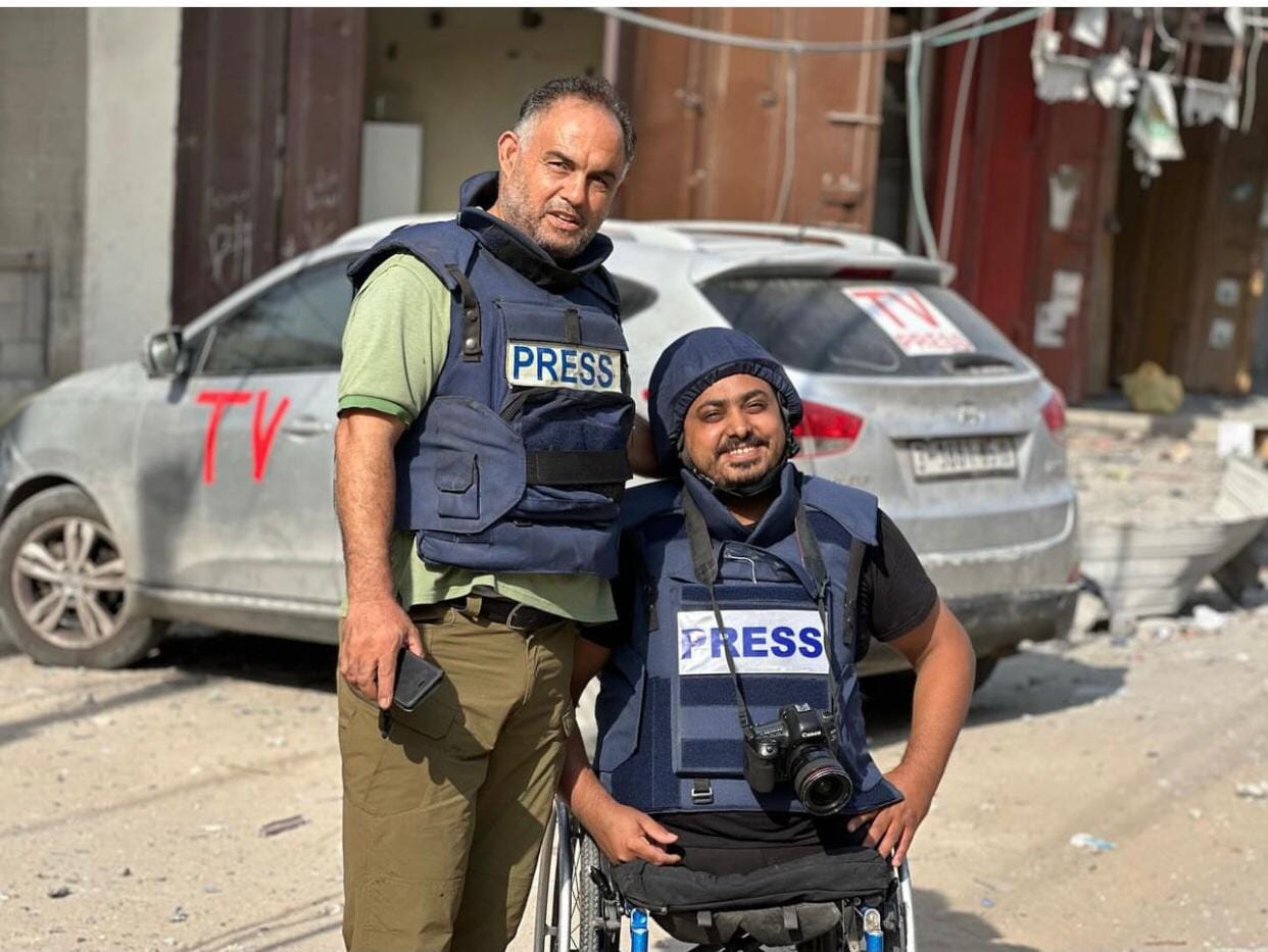 Two men pose for a photo together. On the left - Hatem Moussa. On the right - Momin Faiz. Both are wearing press vests. Behind them, is a press car.