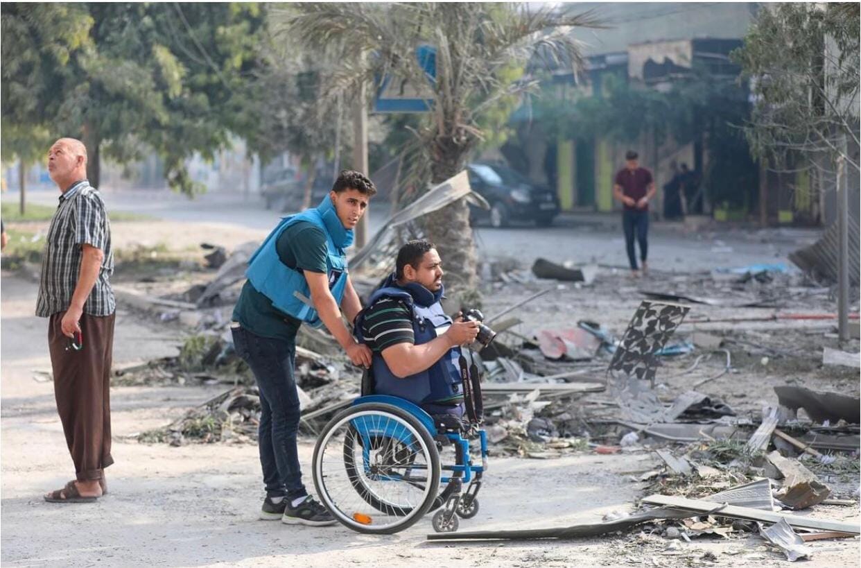 Momin Faiz is in the centre of the photo, taking a photo from his wheelchair. His colleague is pushing him. Both men are wearing press vests and are looking out at the rubble of a destroyed building. 