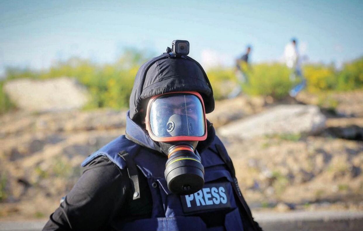 A man wearing a gas mask and a hood with a camera on top looks into the camera. He is in a press vest. Behind him, a landscape is out of focus.  