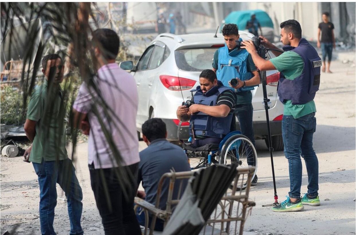 Momin adjusts his equipment with two cameramen behind him. In front of him are three other men, two are standing and one is sitting in a chair.