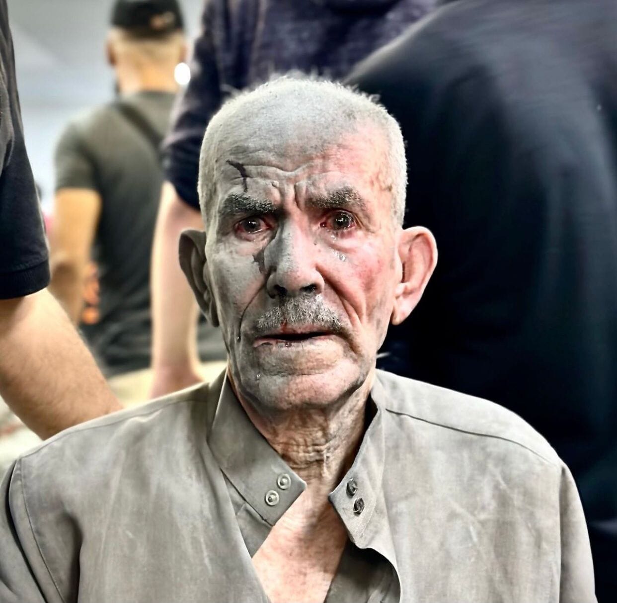 An elderly man, covered in dust looks into the camera. His eyes are bloodshot and filled with tears. He has blood on his face and in his mouth.