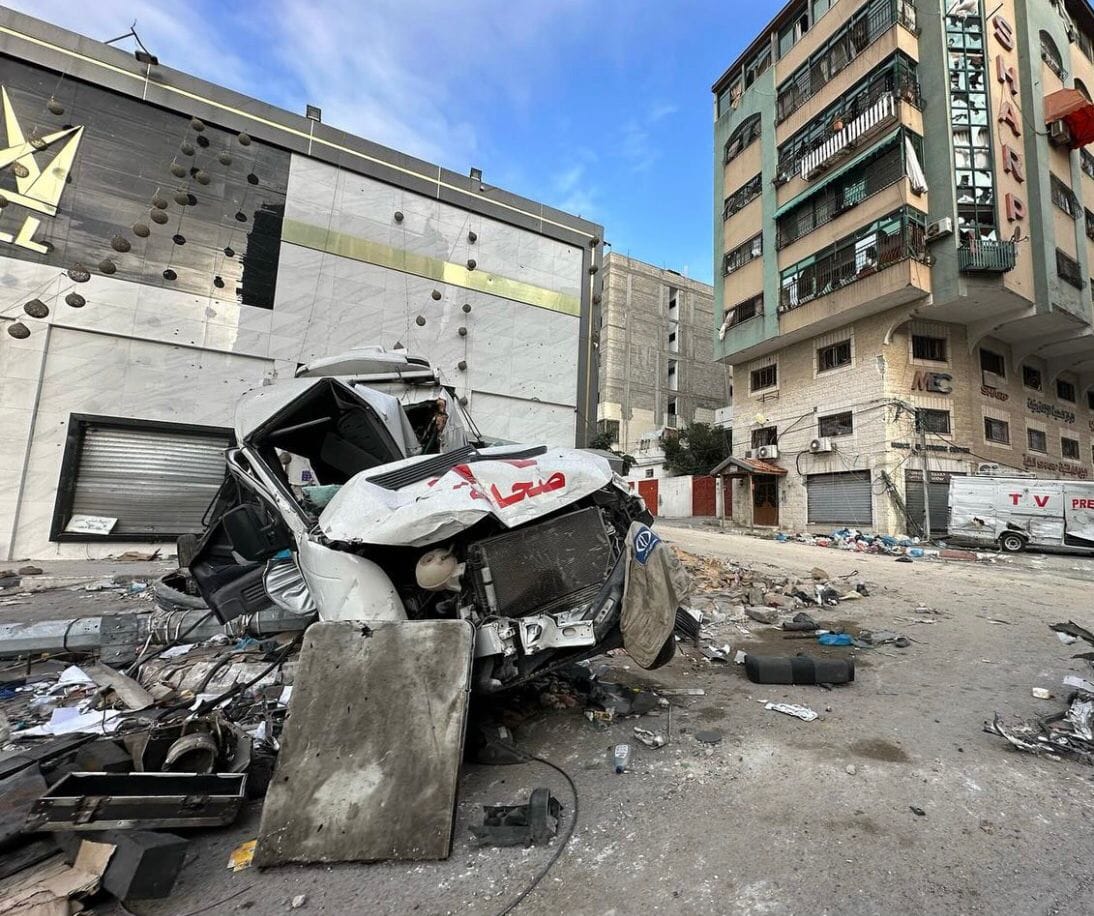 A heavily damaged vehicle sits amongst rubble in the street. The vehicle has red letters on it, indicating that it is a press van.